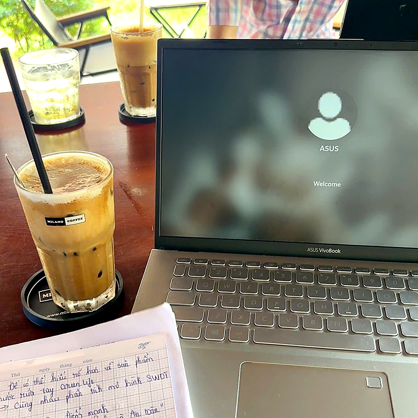 Ly cà phê sữa đá Milano bên laptop và sổ tay trên bàn gỗ